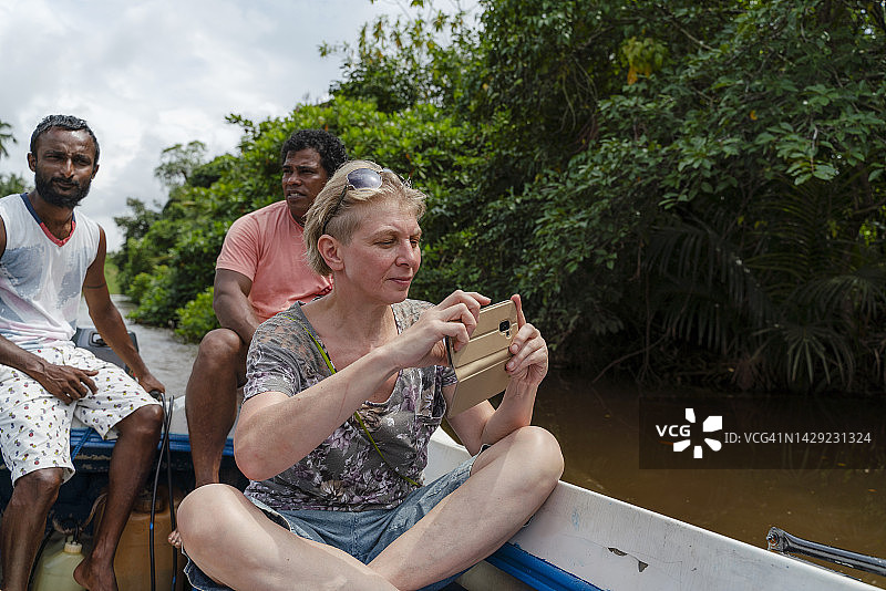 年长的白人短发女游客在斯里兰卡本托塔河乘船旅行，由两位当地导游陪同，用智能手机拍照图片素材