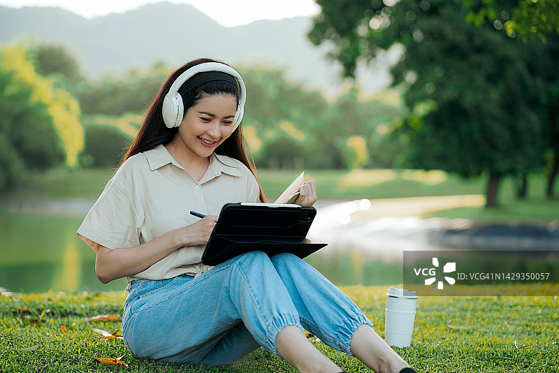 在公园里使用平板电脑和无线耳机上网课的女大学生图片素材
