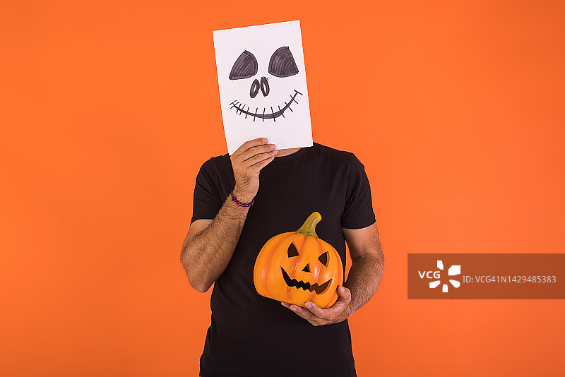 男子用画有恐怖万圣节笑脸的纸遮住脸，手捧南瓜，橙色背景，庆祝亡灵节和狂欢节图片素材