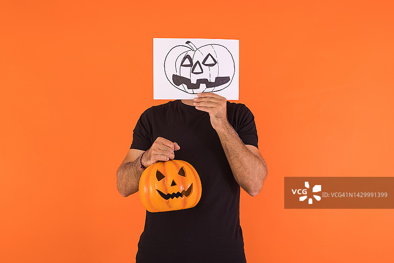 男人用画有万圣节南瓜灯的纸遮住脸，拿着南瓜，橙色背景。庆祝、亡灵节和狂欢节的概念图片素材