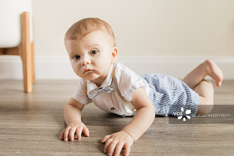 2022年秋季，一个表情严肃的九个月大疫情男婴，光着脚，穿着蓝白条纹短裤和棕褐色吊带，穿着白色短袖衬衫，系着领结，坐在家里地板上图片素材