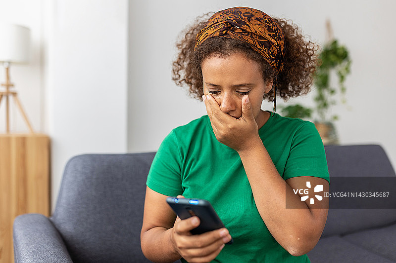 沮丧的年轻成年女子在智能手机上阅读信息。心烦意乱的青少年女学生坐在沙发上，手持手机，手捂着脸。坏消息、负面情绪、糟糕的感觉概念图片素材