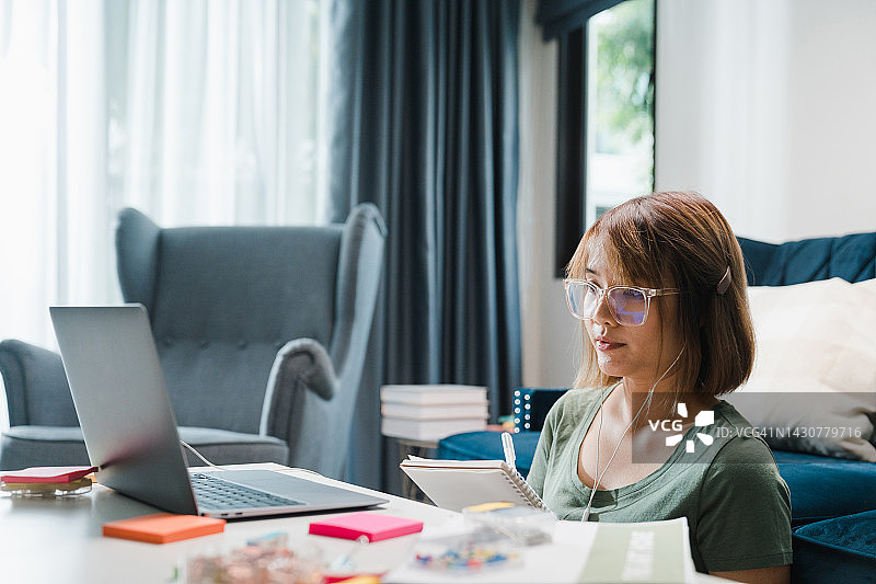 年轻女孩戴着耳机在家用笔记本电脑在线学习并写笔记图片素材