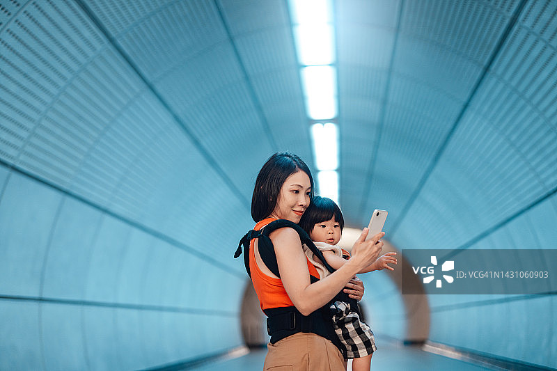 年轻的母亲用婴儿背带背着女儿，在未来主义隧道中使用智能手机图片素材