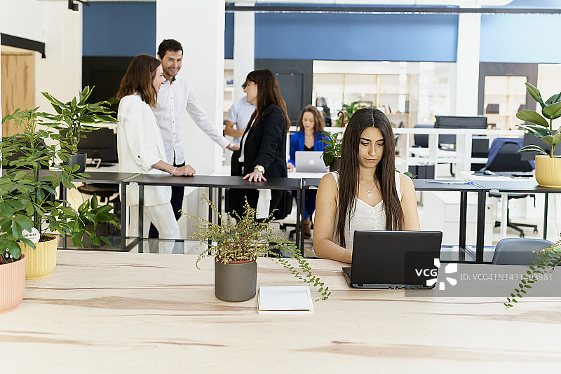 在装饰精美的办公室里，年轻女人坐在笔记本电脑前工作，同事们在她身后高兴地聊天图片素材