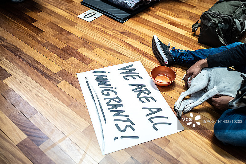 难民男子和他的狗坐在地上，靠近写着“我们都是移民！”的标志图片素材