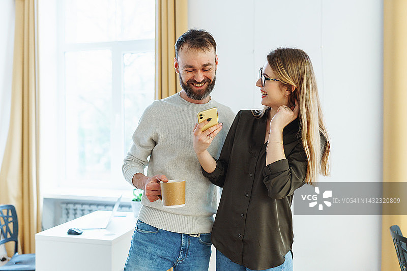 办公室恋情。微笑的男人和女人一起聊天，喝咖啡，并通过手机分享新闻图片素材