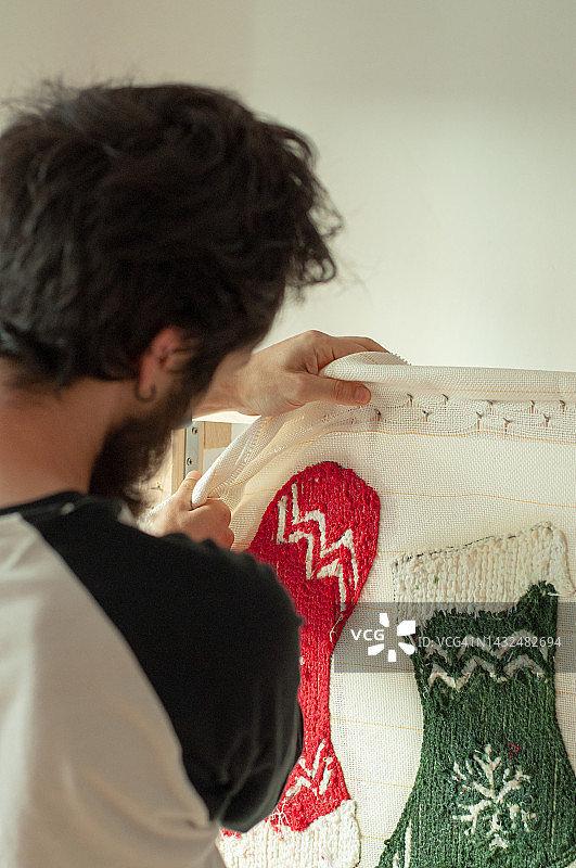 男人用手从框架上取下有机棉布，制作簇绒地毯，圣诞图案是红绿相间的圣诞袜和雪花，竖版构图，自然光图片素材