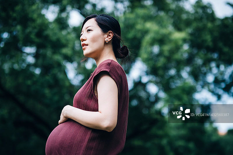 年轻的亚洲孕妇在公园里抚摸着隆起的肚子，放松身心，呼吸着新鲜空气，享受着大自然的宁静。准妈妈期待着宝宝的到来，爱和联结，积极健康的怀孕生活方式图片素材