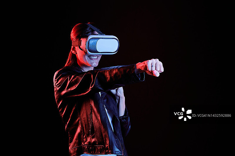 年轻白人女子戴VR眼镜在黑背景下挥拳，红蓝光照亮图片素材