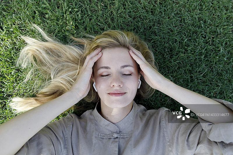 躺在草地上用无线入耳式耳机听音乐的微笑女人图片素材