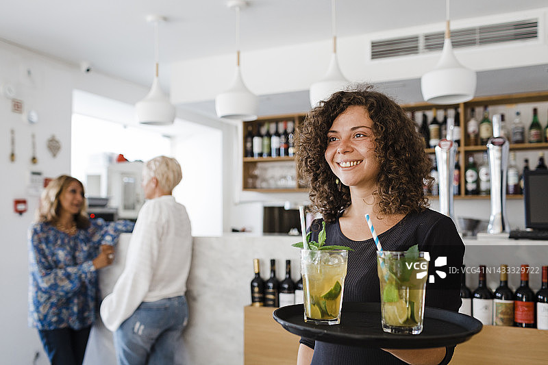 快乐的年轻女服务员在餐厅托盘上端着鸡尾酒图片素材