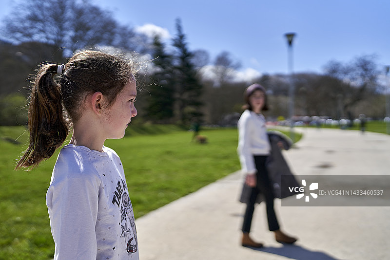 阳光明媚的日子里，一位棕色头发的女孩站在公园的人行道上，凝视着走远的母亲和哥哥图片素材