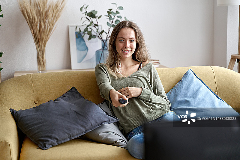 年轻女人在家里的沙发上看电视，面带微笑图片素材