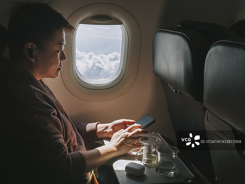 中国女性在飞机上坐在乘客窗口座位上使用智能手机图片素材