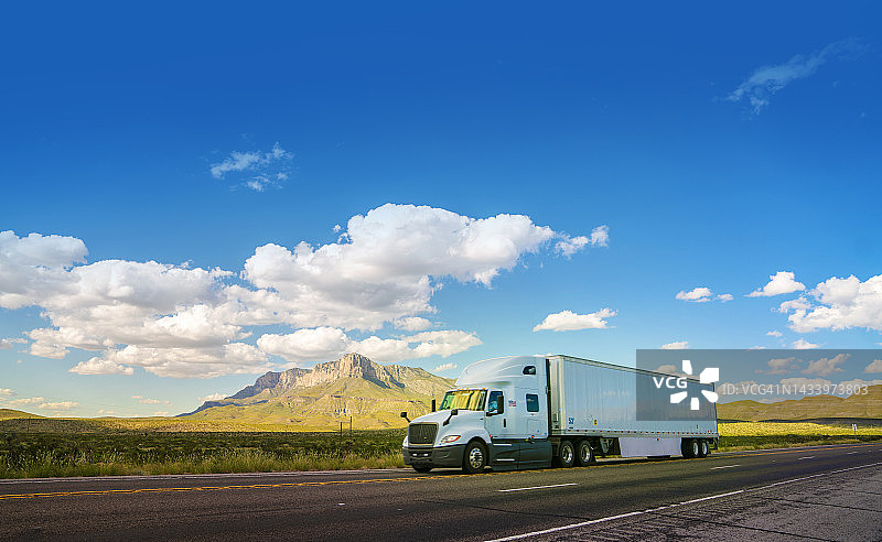 在德克萨斯州乡村公路上行驶的白色半挂卡车，背景是埃尔卡皮坦山图片素材
