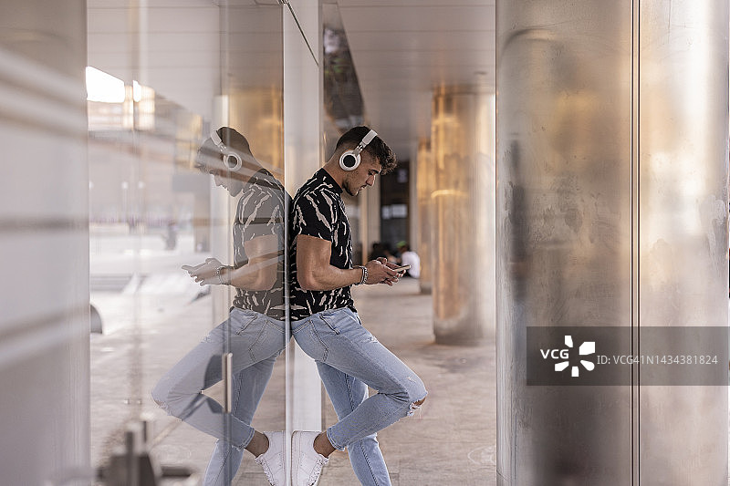 城市中倚靠玻璃墙的男人，用手机发信息听音乐，现代城市生活概念图片素材
