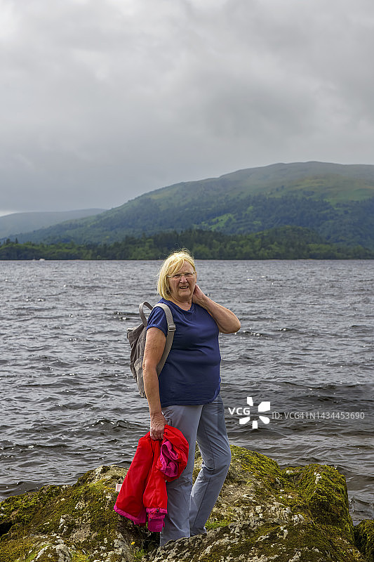 活跃的老年女性徒步旅行者站在英国苏格兰卡舍尔附近的洛蒙德湖岸边的露头上看着相机。图片素材