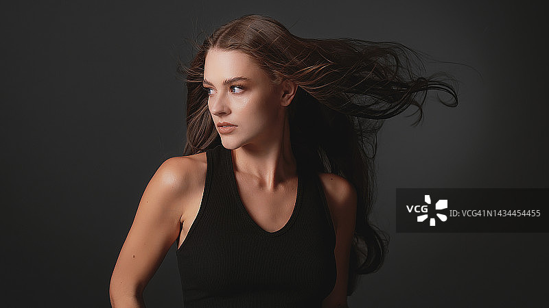 深色背景下迷人的棕发美女：闪亮健康的棕色头发随风飘动图片素材
