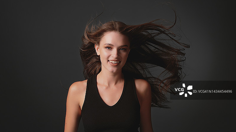 深色背景下迷人的棕发美女：闪亮健康的棕色头发随风飘动图片素材
