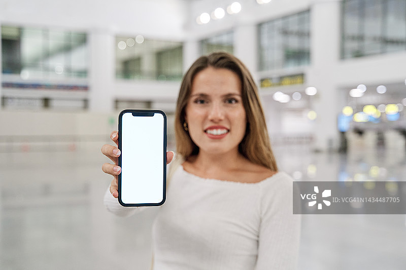在机场或火车站航站楼里，一位美丽的微笑的年轻白人女性的正面肖像，向相机展示一部带有空白屏幕的手机图片素材