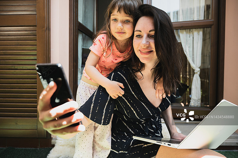在家工作的母亲拥抱她3岁的孩子，同时使用手机图片素材