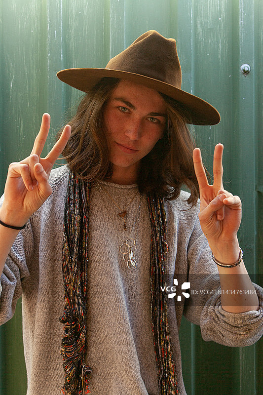 英俊的年轻男子对着镜头做出“和平”手势，戴着宽边帽，看起来自信而放松图片素材