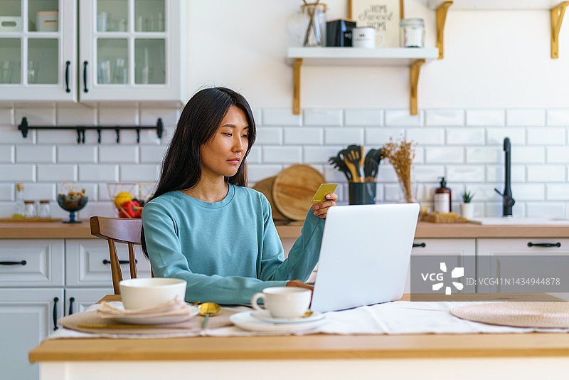 在线购物概念。亚洲女性坐在厨房，用信用卡支付购物费用，计划旅行，选择度假，寻找机票，预订酒店图片素材