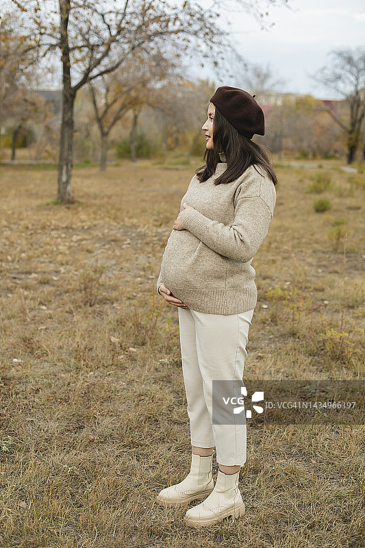 穿着米色裤子和米色毛衣，戴着贝雷帽的女人在秋日里将手放在肚子上图片素材