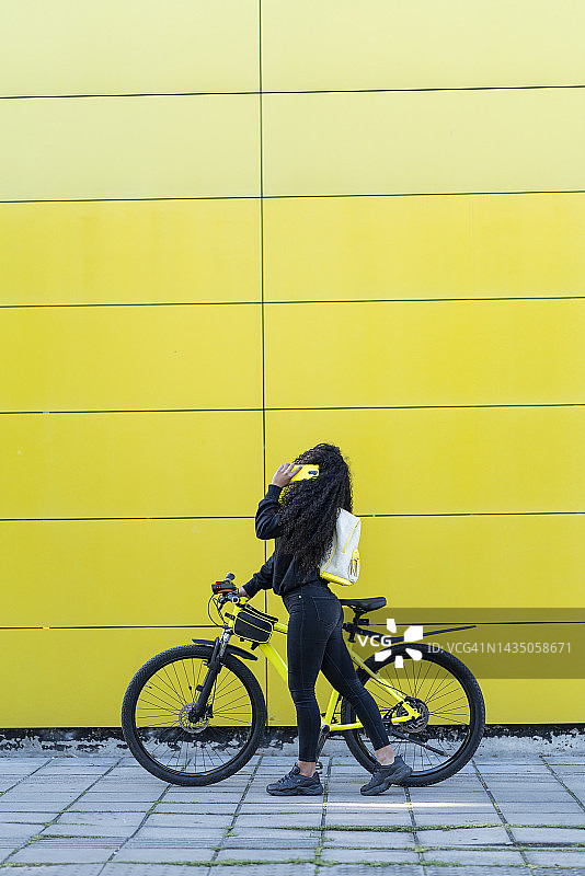 在城市街道上和她的黄色自行车停在黄色墙壁旁的非裔拉丁裔女子图片素材