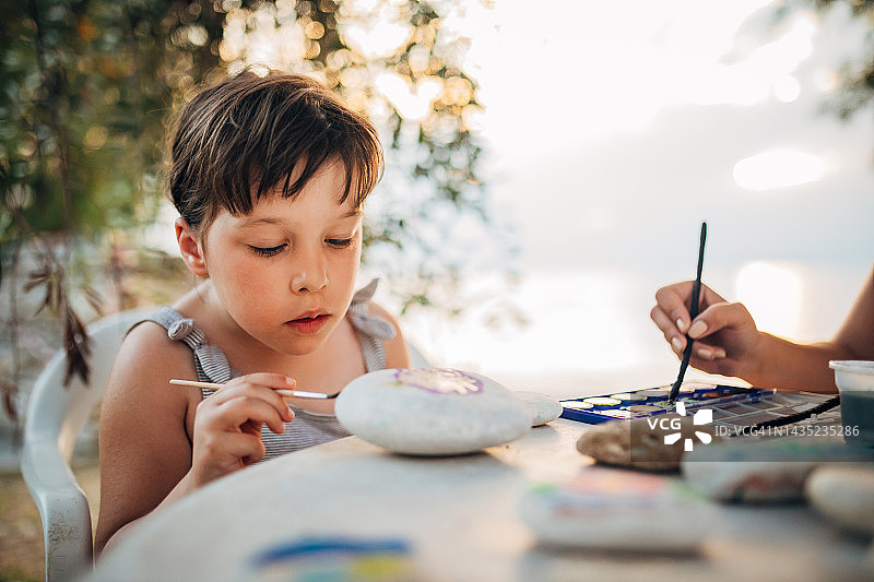 日落时分坐在海滩附近，在岩石上绘画的可爱小女孩图片素材