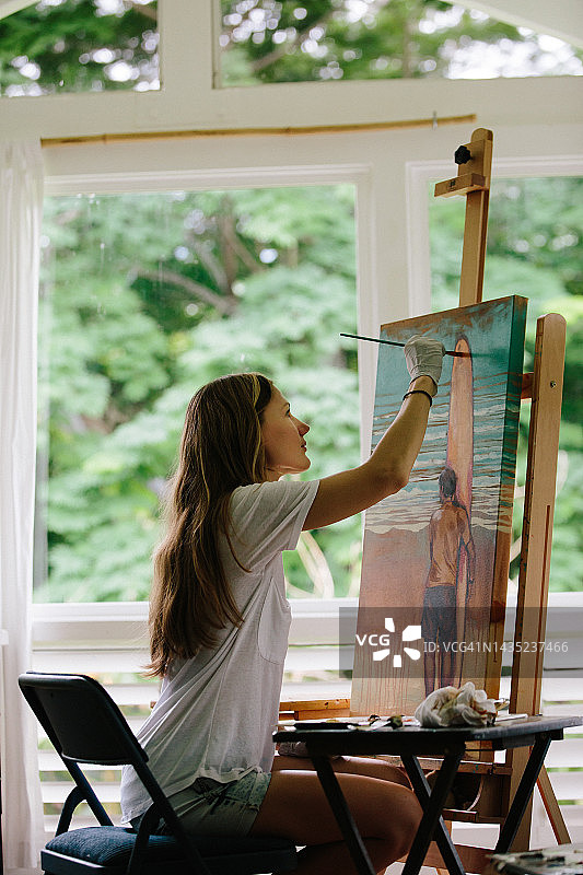 年轻女子在宽敞明亮的工作室里用白色T恤在画布上绘画，可欣赏绿色植物的景色。冲浪者站在海边的画作。图片素材