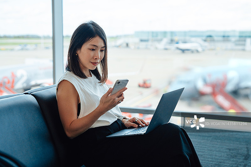 在机场航站楼等候登机时，使用笔记本电脑和智能手机工作的自信年轻女商人图片素材