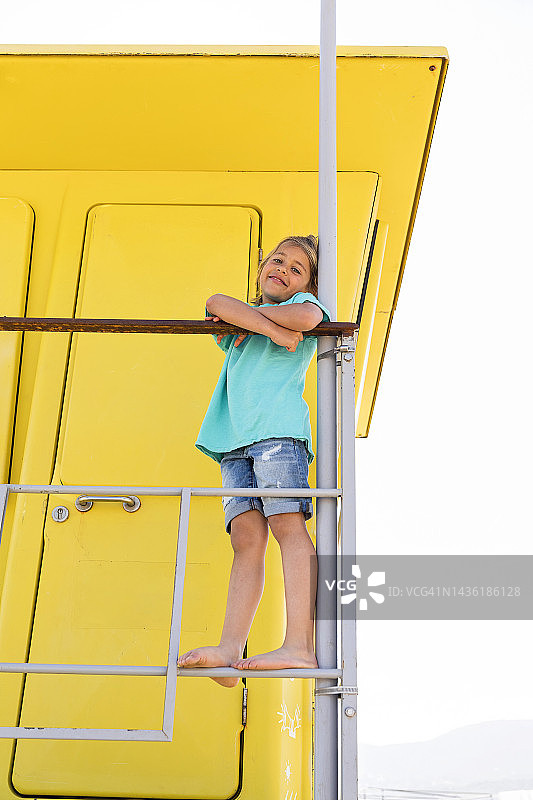 微笑的男孩双臂交叉站在黄色救生员小屋前的栏杆上图片素材