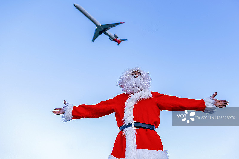 男子身穿圣诞老人服装，在飞行飞机下张开双臂站立图片素材