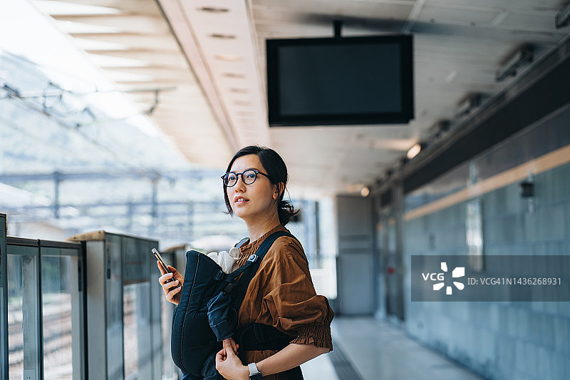 年轻的亚裔母亲手持智能手机，用婴儿背带带着女儿在城市里乘坐地铁图片素材