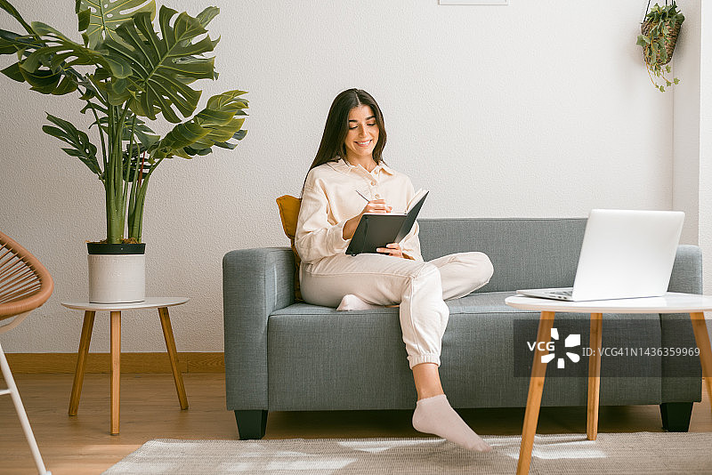 正面视角：一位快乐的年轻女子，留着长长的黑发，穿着舒适的衣服，无忧无虑地微笑，一边在笔记本或日记本上写作，一边坐在她家的客厅的沙发上。图片素材