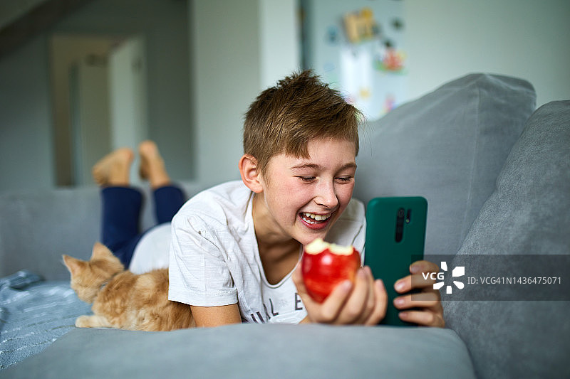 微笑的金发小男孩吃苹果并使用智能手机图片素材