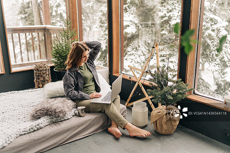 年轻漂亮的亚洲女性在房间的床上使用笔记本电脑，房间里有圣诞树，她有着深色的长发，穿着舒适的白色针织毛衣。无忧无虑的快乐少女学生在圣诞节的房子里的肖像图片素材