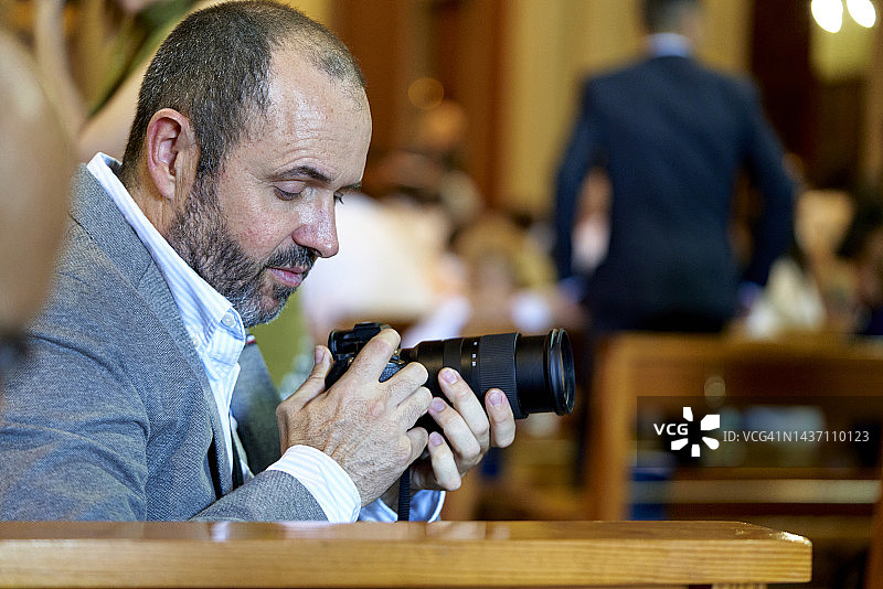 一位灰头发西装的老年男人坐在教堂里拿着相机，背景中有几个人图片素材