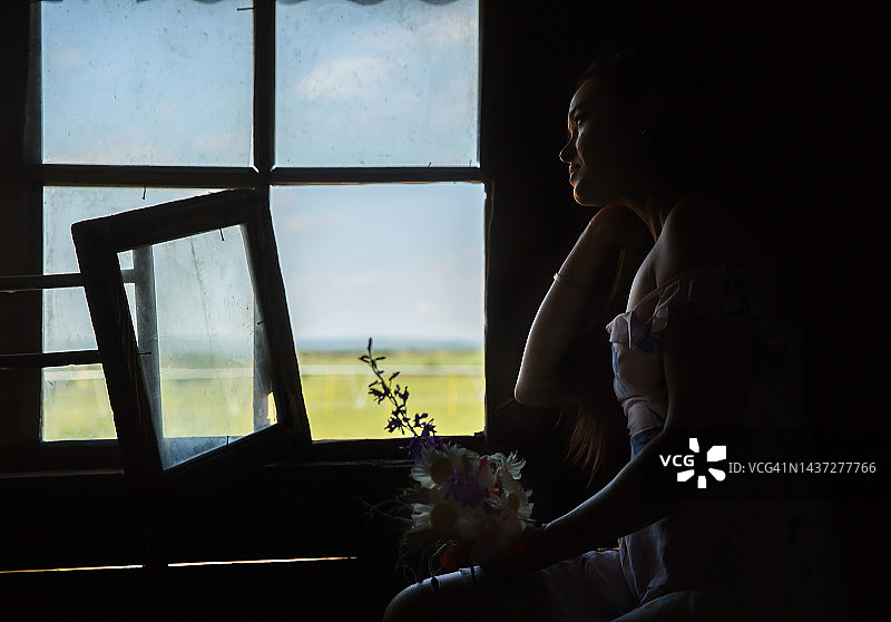年轻漂亮的女子身穿白色夏装，坐在黑暗的房子里，靠近鲜花盛开的窗户图片素材