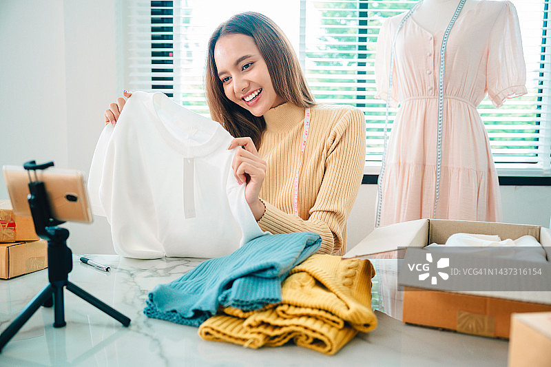 亚洲美女在家直播销售布料产品。年轻女子使用手机视频通话向顾客展示商品并介绍细节。远程购买和采购购物概念图片素材