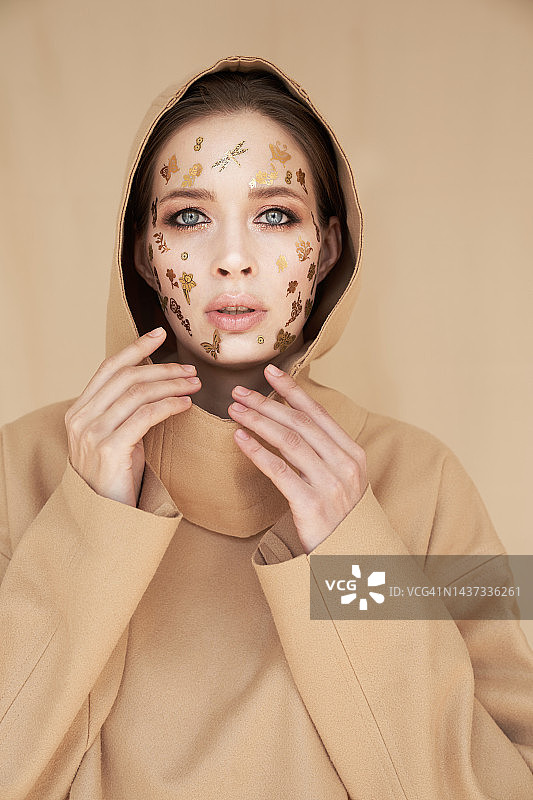 戴着头巾、身穿米色西装的年轻女子站在米色背景前，脸上贴着金色装饰贴纸。伊斯兰女性、万圣节派对、美容、化妆概念图片素材