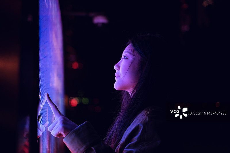 年轻的亚洲女性在夜晚的金融区使用触摸屏图片素材