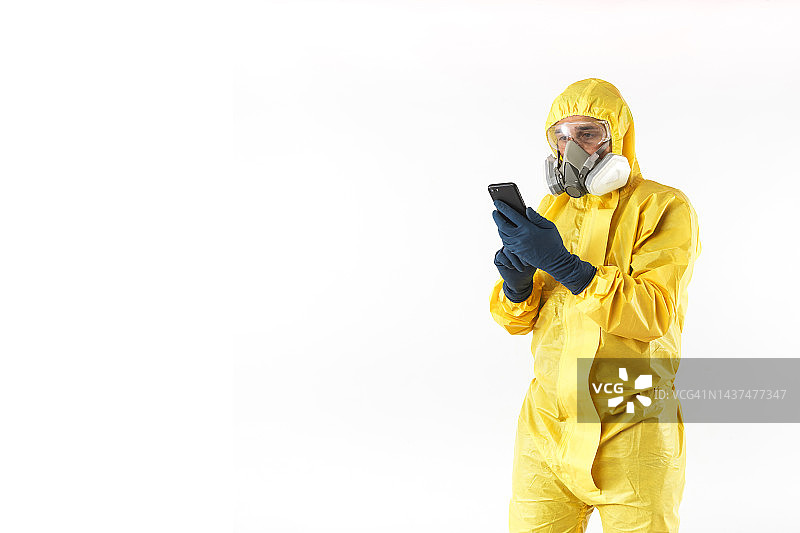 身穿黄色核防护服、戴着面罩和防护眼镜的技术员在白色背景下查看手机。核能与疫情概念图片素材