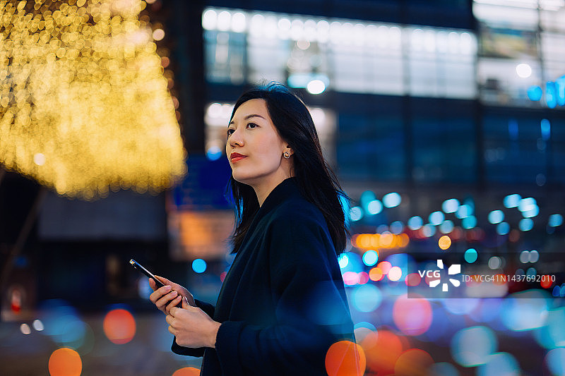 年轻的亚洲女性在城市街道上使用手机App预约出租车，背景是灯火通明的城市夜景和彩色散景。生活方式与交通科技，便捷且值得信赖的汽车服务图片素材