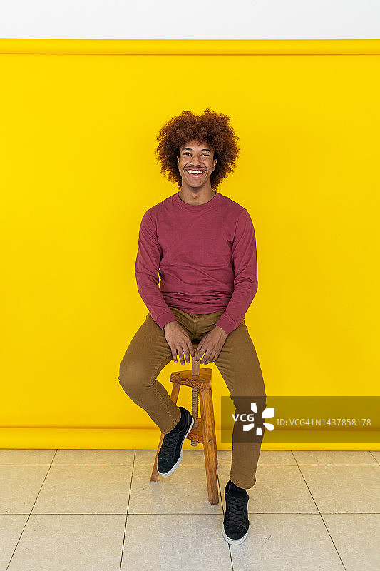 快乐的黑人时髦青年男子坐在黄色背景的摄影棚里对着镜头微笑的竖版照片图片素材