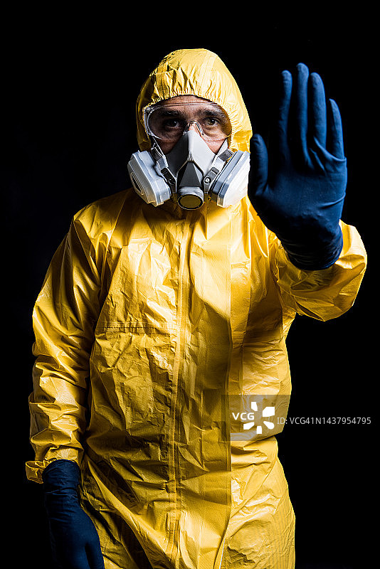 身穿黄色核防护服、戴面罩和防护眼镜的技术员在黑色背景下用手势示意停止，核能与疫情概念图片素材