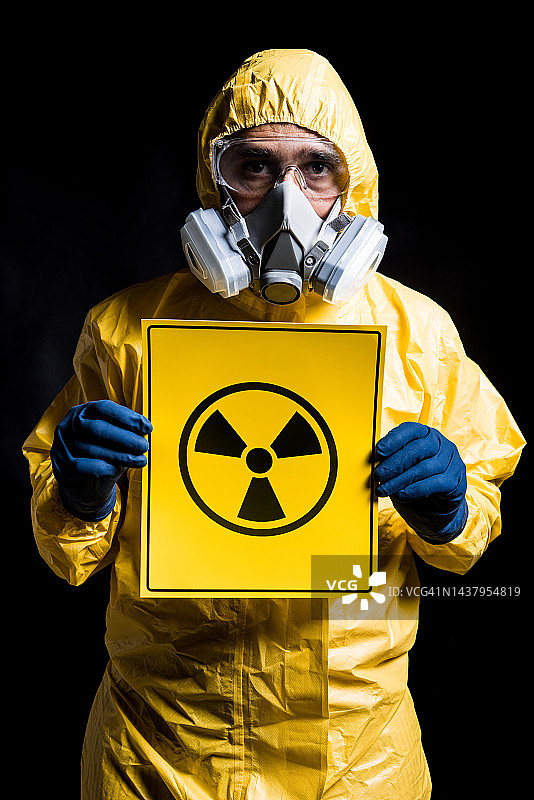 身穿黄色核防护服、戴着面罩和防护眼镜的技术员肖像，手持带有核标志的标志，黑色背景。核能与疫情概念。图片素材