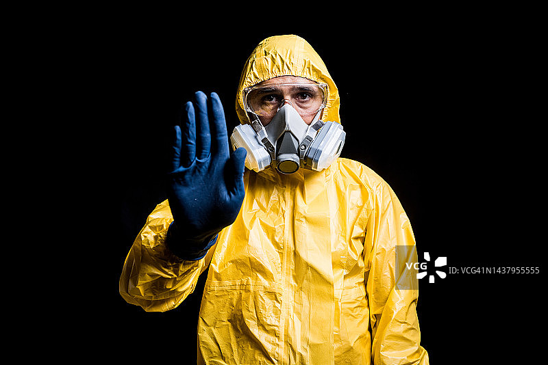 身穿黄色核防护服、戴面罩和防护眼镜的技术员在黑色背景下用手势示意停止，核能与疫情概念图片素材